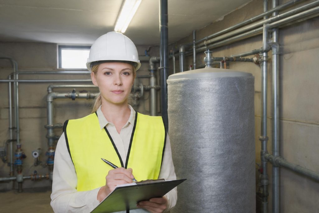 Legionella risk assessor writing on a clipboard beside a hot water cylinder in a plant room